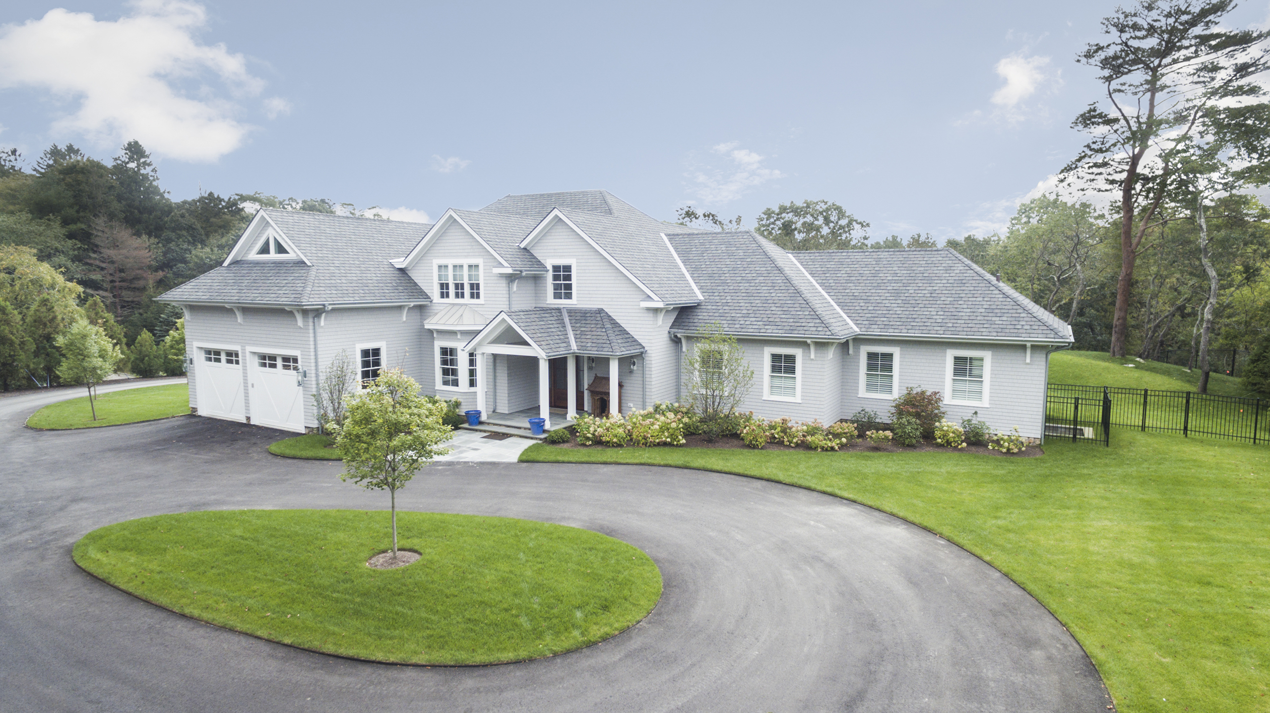 A large white house with a gray roof, surrounded by a curved driveway and well-maintained lawns, set against a backdrop of trees and a cloudy sky.