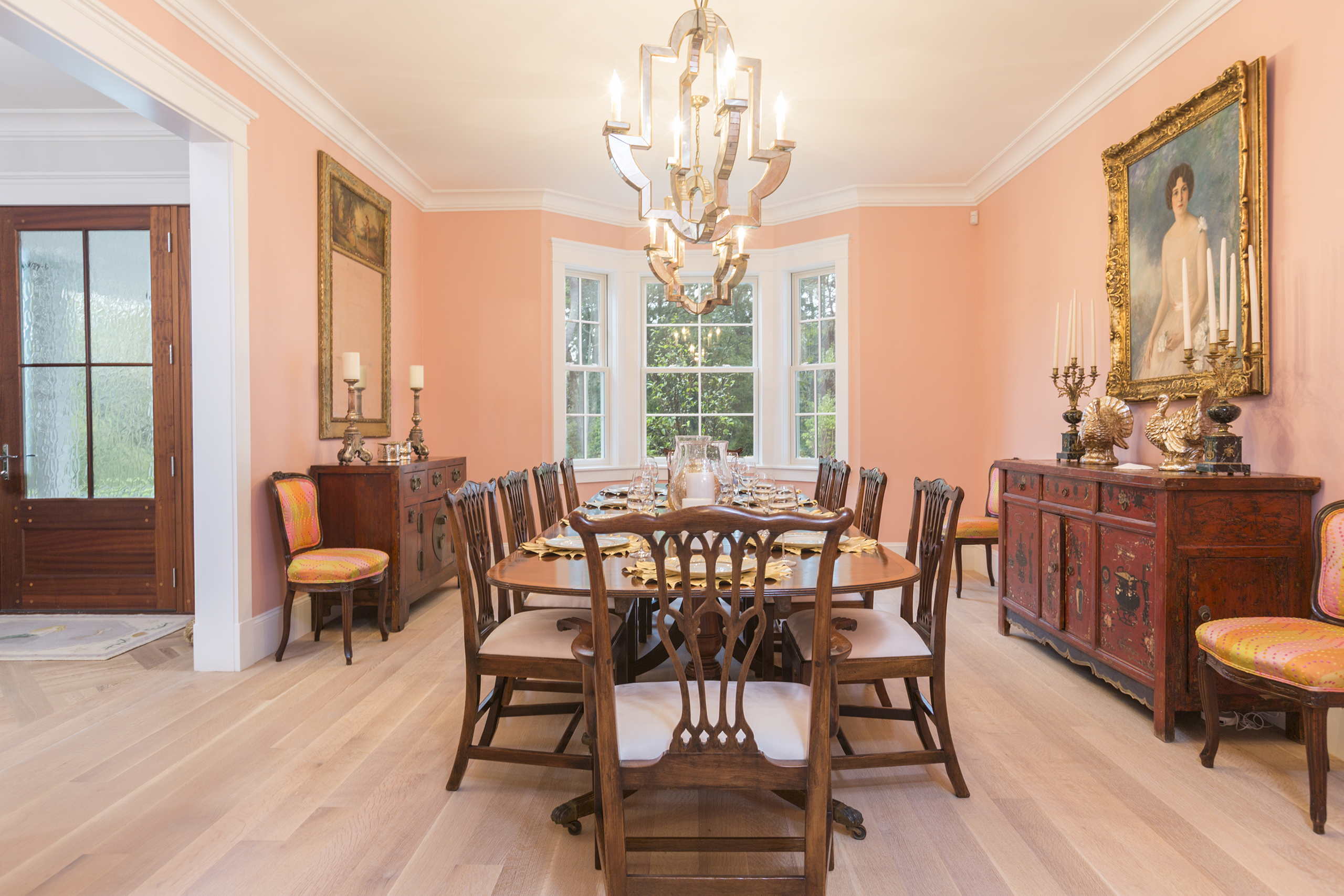 A dining room with a large wooden table, ten chairs, a chandelier, and two sideboards. Walls are pink with artwork. Large window at the back.