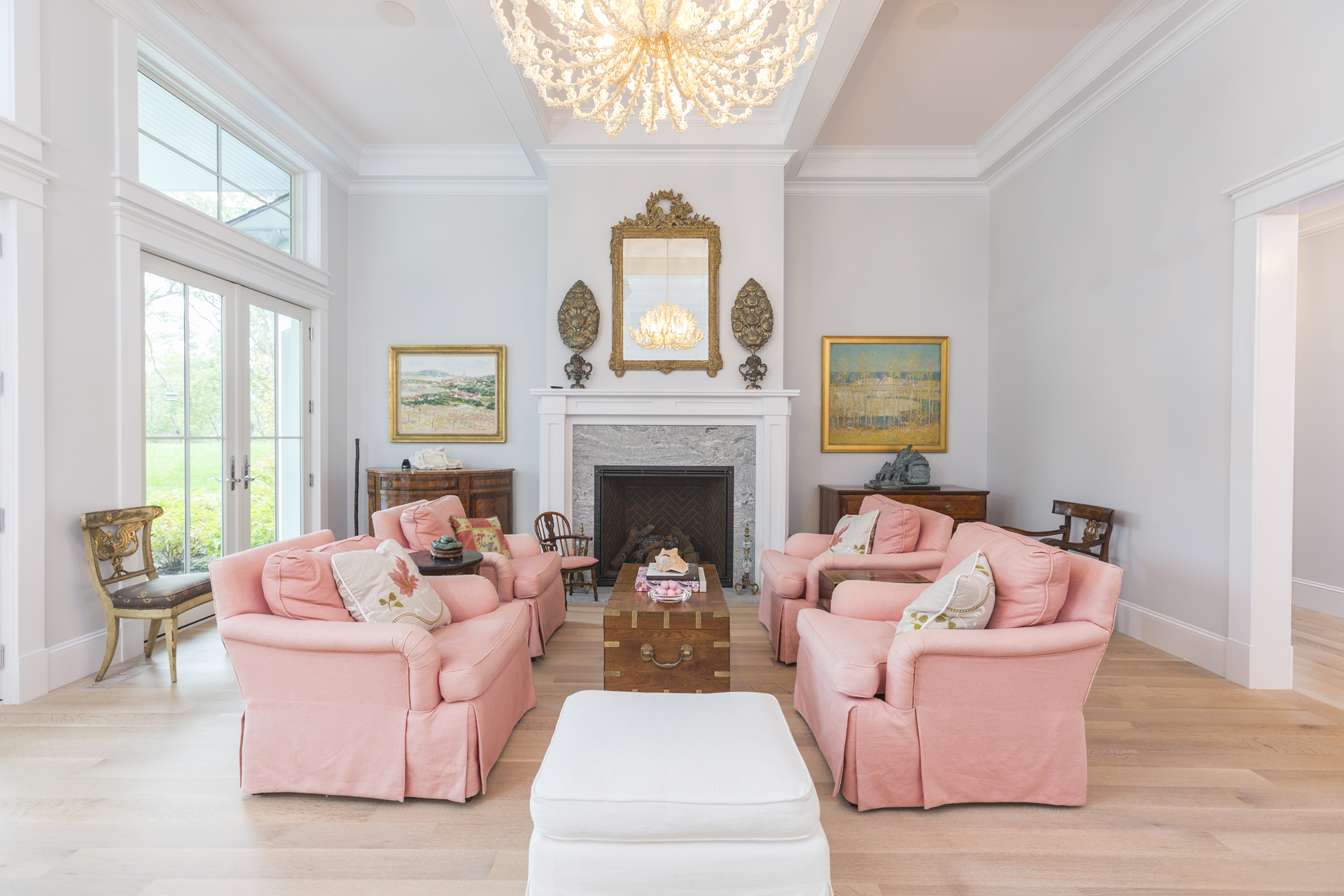 Elegant living room with pink armchairs, chandelier, fireplace, and wall art. Large windows provide a view outside. A white ottoman and a wood table are in the center.