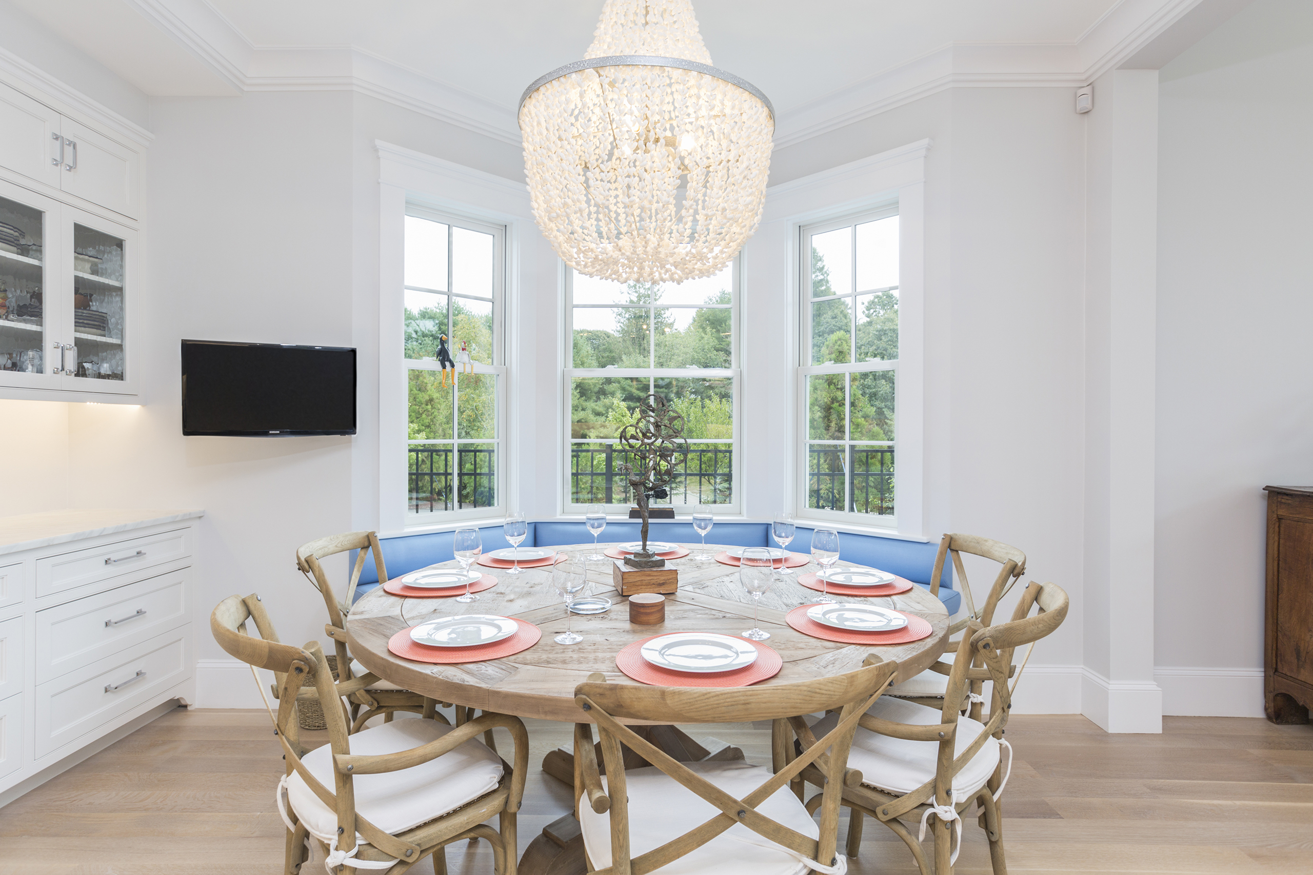 Dining room with large round wooden table, surrounded by six chairs with white cushions. Table is set with plates and red chargers. A crystal chandelier hangs above. Three windows in the background.