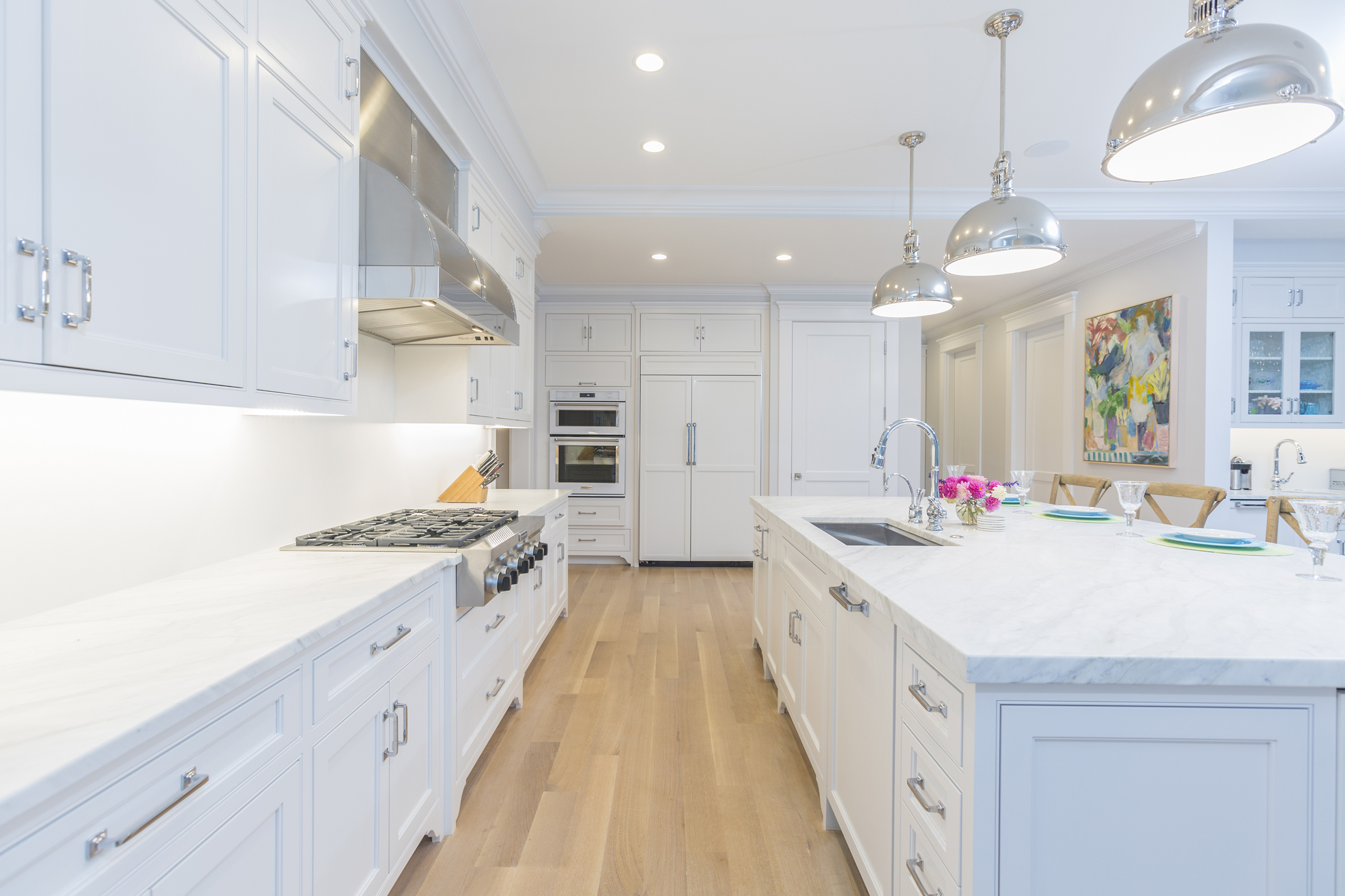 A spacious, modern kitchen with white cabinets, marble countertops, wooden floor, stainless steel appliances, and pendant lights. A colorful painting hangs on the wall.