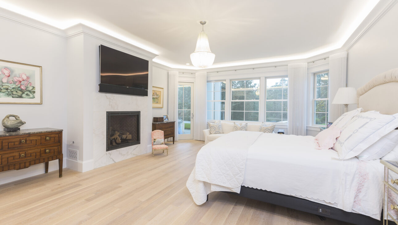A spacious bedroom with a bed, fireplace, wall-mounted TV, and large windows. Light wooden flooring and a small dresser are also visible.