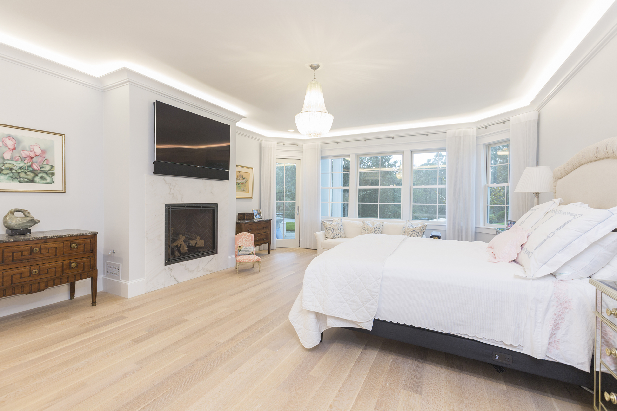 A spacious bedroom with a bed, fireplace, wall-mounted TV, and large windows. Light wooden flooring and a small dresser are also visible.