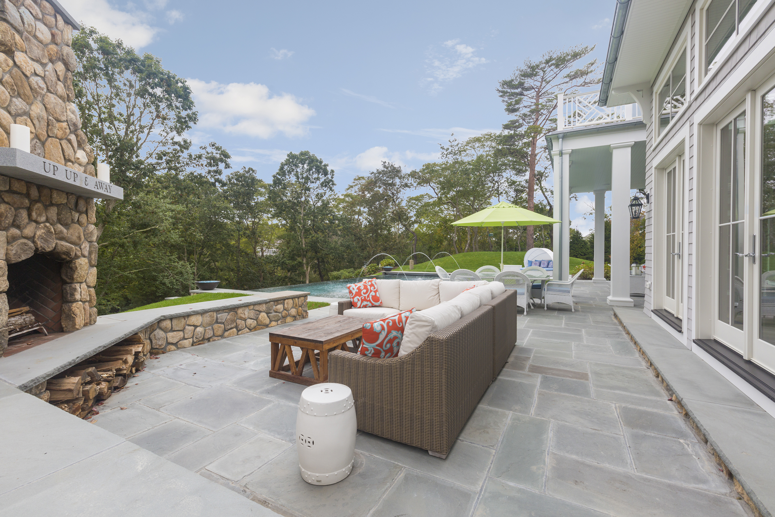 Patio with a wicker sofa, stone fireplace, and pool. Green umbrella over a dining table. Trees and cloudy sky in the background.
