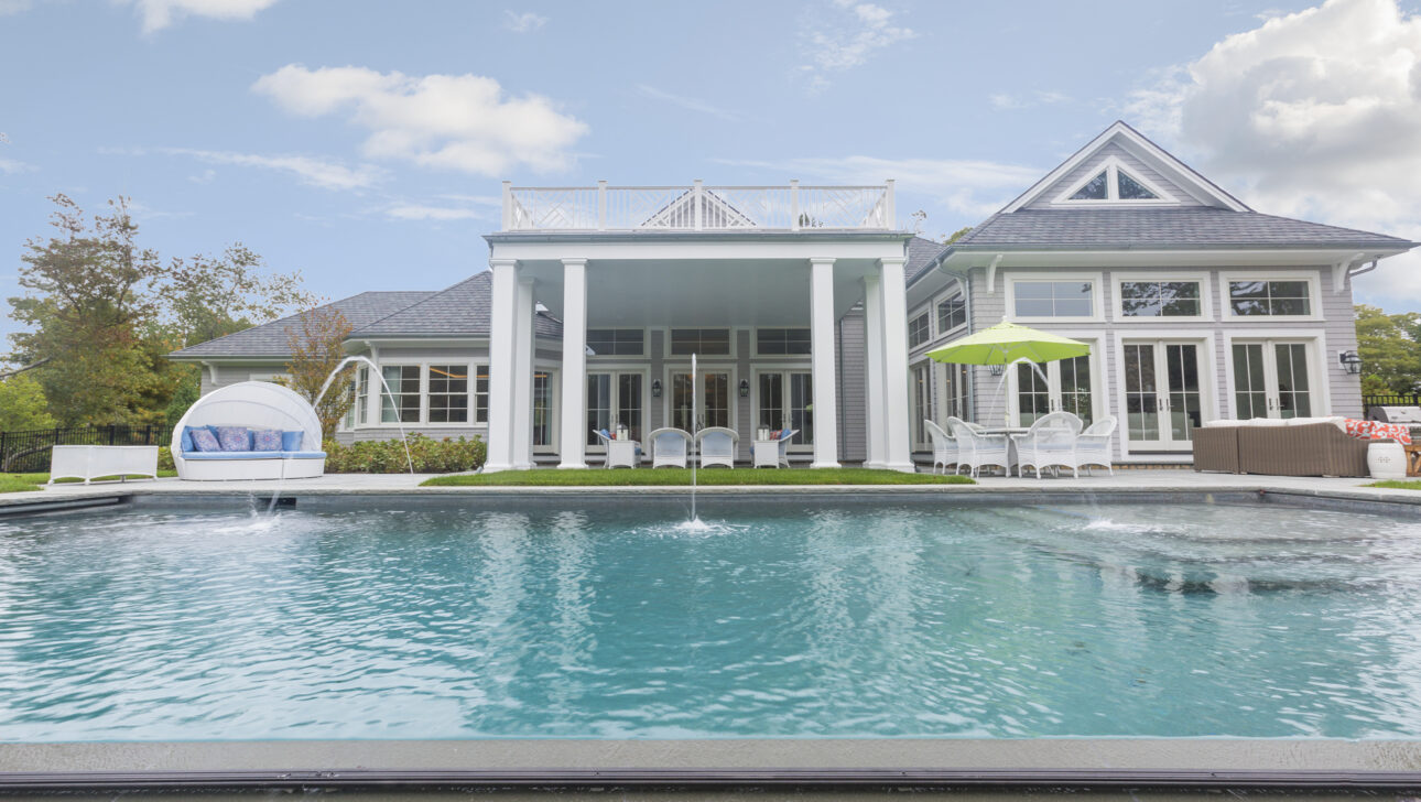 A luxurious house with a large swimming pool, white patio furniture, and a green umbrella in the backyard under a partly cloudy sky.