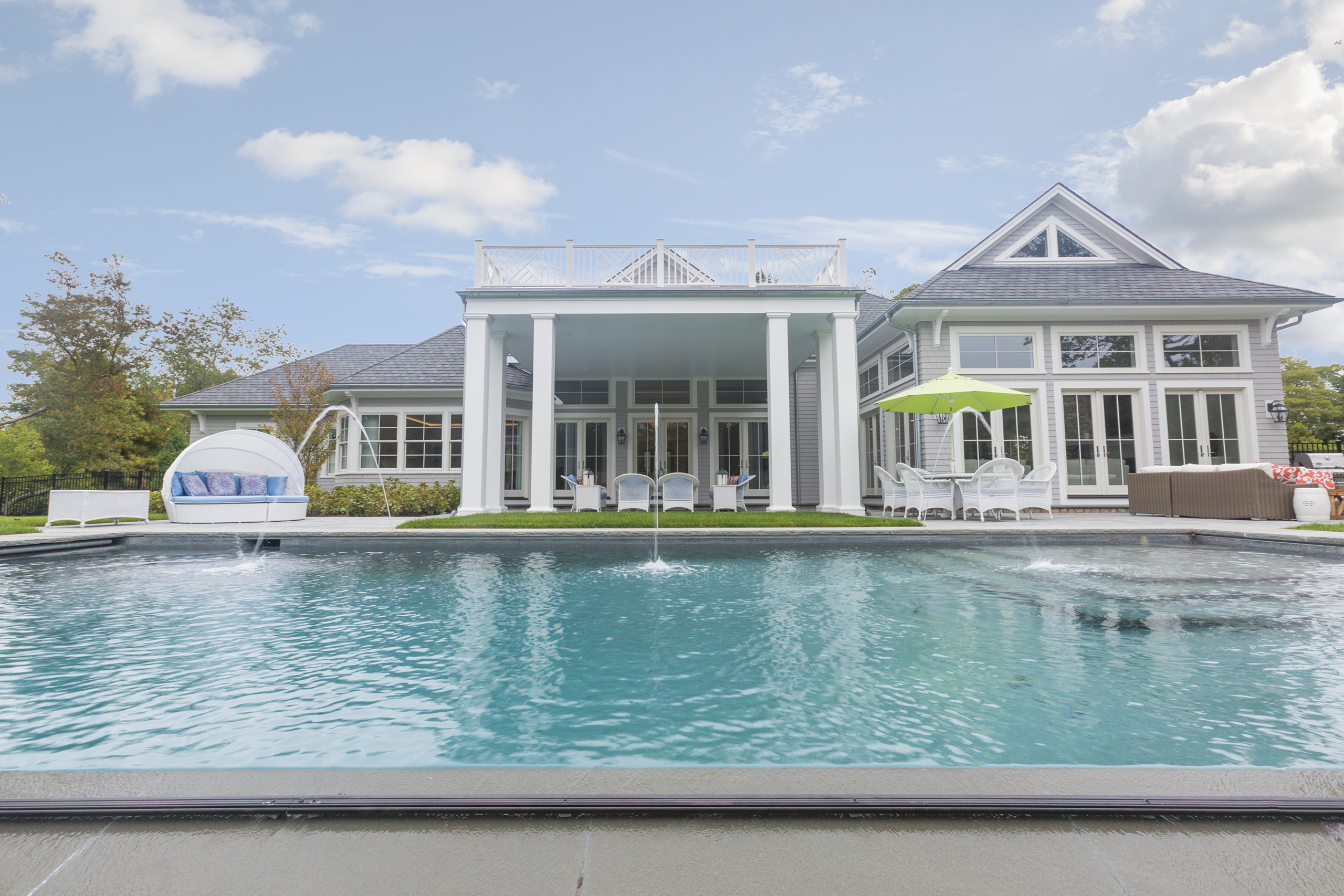 A luxurious house with a large swimming pool, white patio furniture, and a green umbrella in the backyard under a partly cloudy sky.