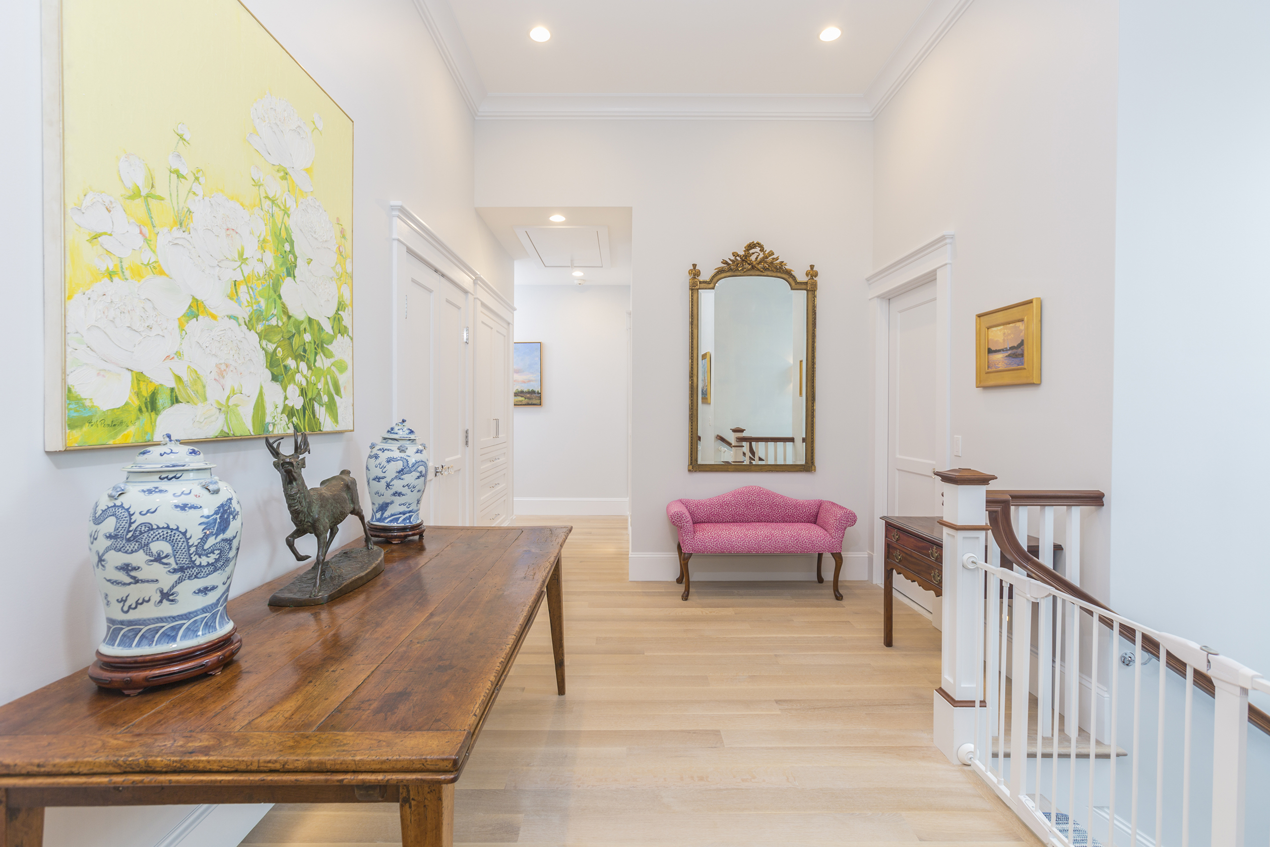 A hallway with a wooden table, decorative vases, a mirror, a pink bench, and a painting of white flowers on the wall. Wooden flooring and white walls complete the setting.