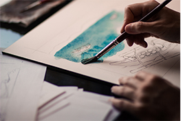 A person uses a paintbrush to apply teal watercolor on paper, surrounded by sketch outlines and additional sheets.