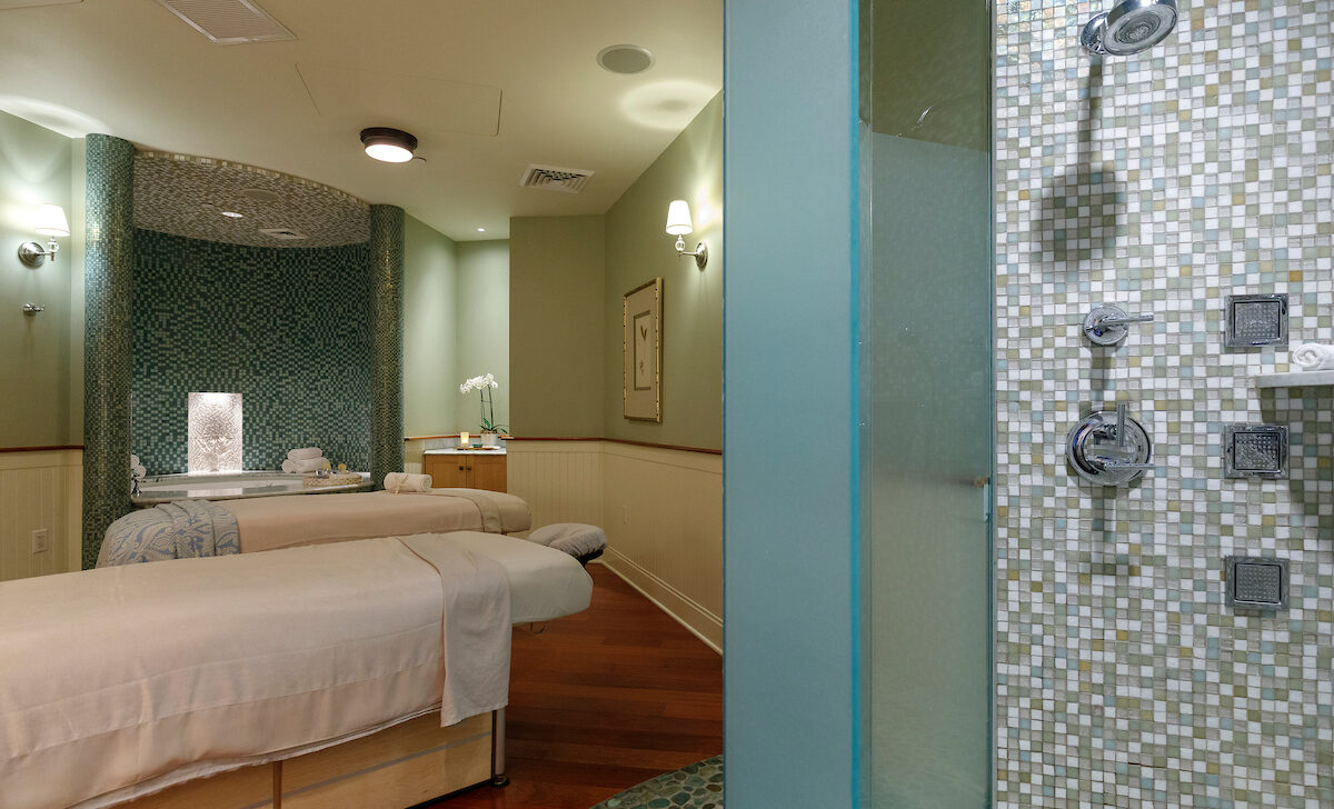 A spa room with two massage tables has soft lighting, green walls, and a tiled shower area with multiple showerheads. A small table holds towels and a flower.