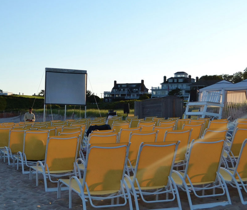 Rows of yellow lounge chairs face a projection screen on a sandy beach with houses and clear sky in the background.