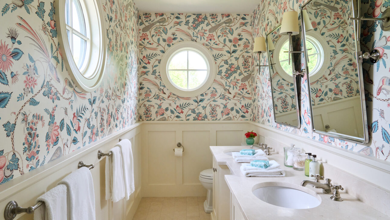 A bathroom with floral wallpaper, a round window, a mirror, two wall lights, a sink with toiletries, and white towels on a rack.