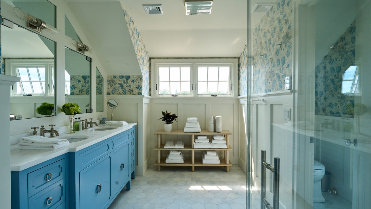 Bright bathroom with blue double vanity, large mirrors, and hexagonal floor tiles. A wooden shelf holds towels and a plant. A glass shower is on the right, and two windows provide natural light.
