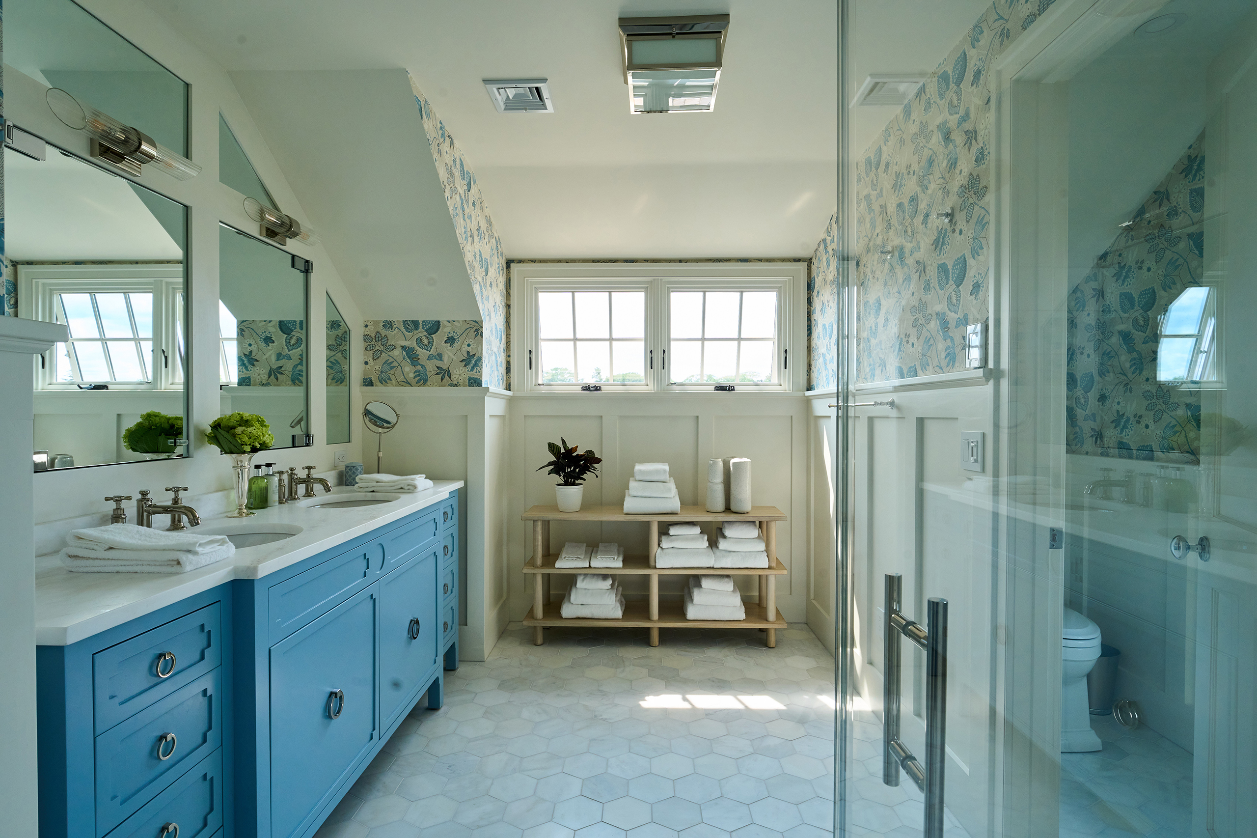 Bright bathroom with blue double vanity, large mirrors, and hexagonal floor tiles. A wooden shelf holds towels and a plant. A glass shower is on the right, and two windows provide natural light.