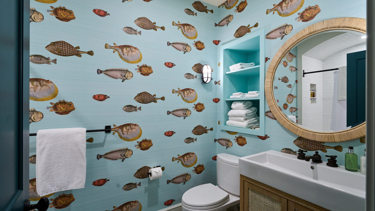 Bathroom with aquatic-themed wallpaper, round mirror, and white fixtures. Shelves hold folded towels.