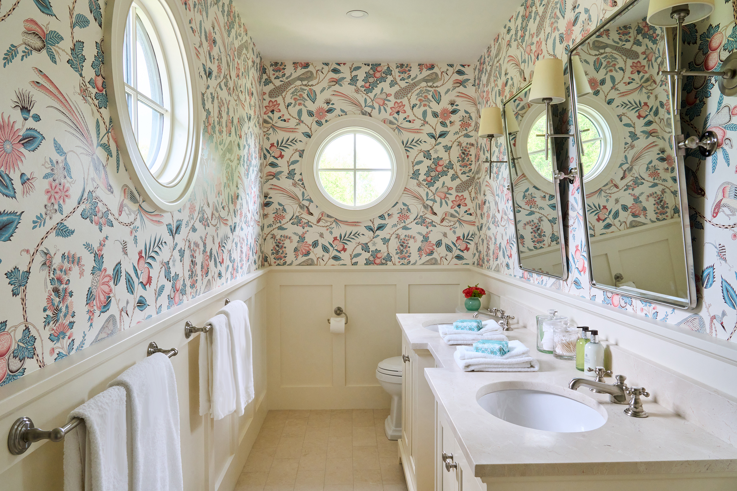 A bathroom with floral wallpaper, a round window, a mirror, two wall lights, a sink with toiletries, and white towels on a rack.