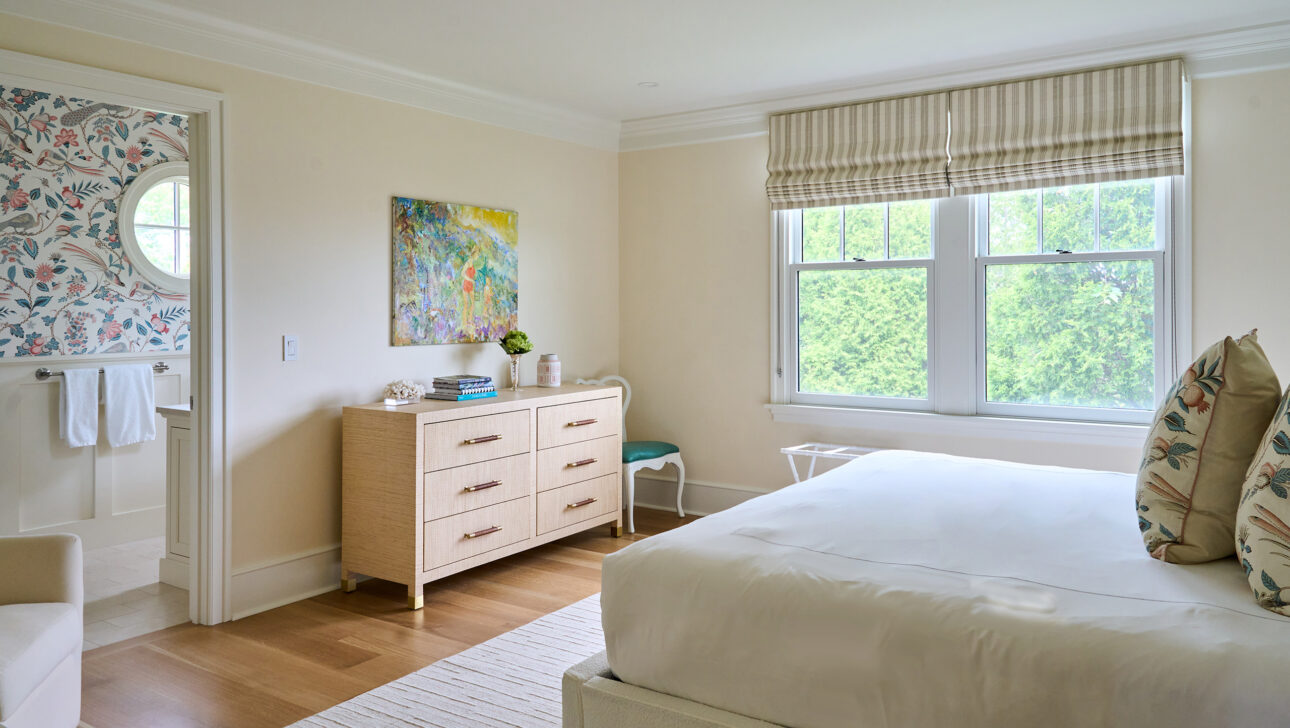 Bedroom with a white bed, beige dresser, artwork, and striped blinds. A bathroom with floral wallpaper is visible through the open door.