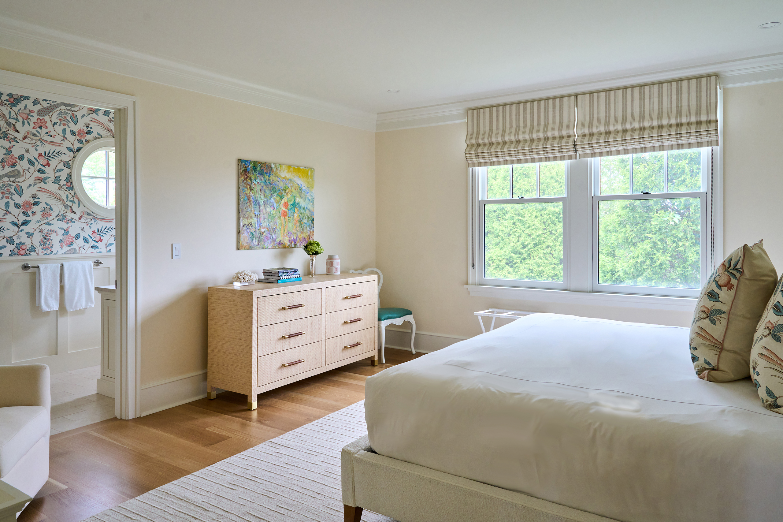 Bedroom with a white bed, beige dresser, artwork, and striped blinds. A bathroom with floral wallpaper is visible through the open door.