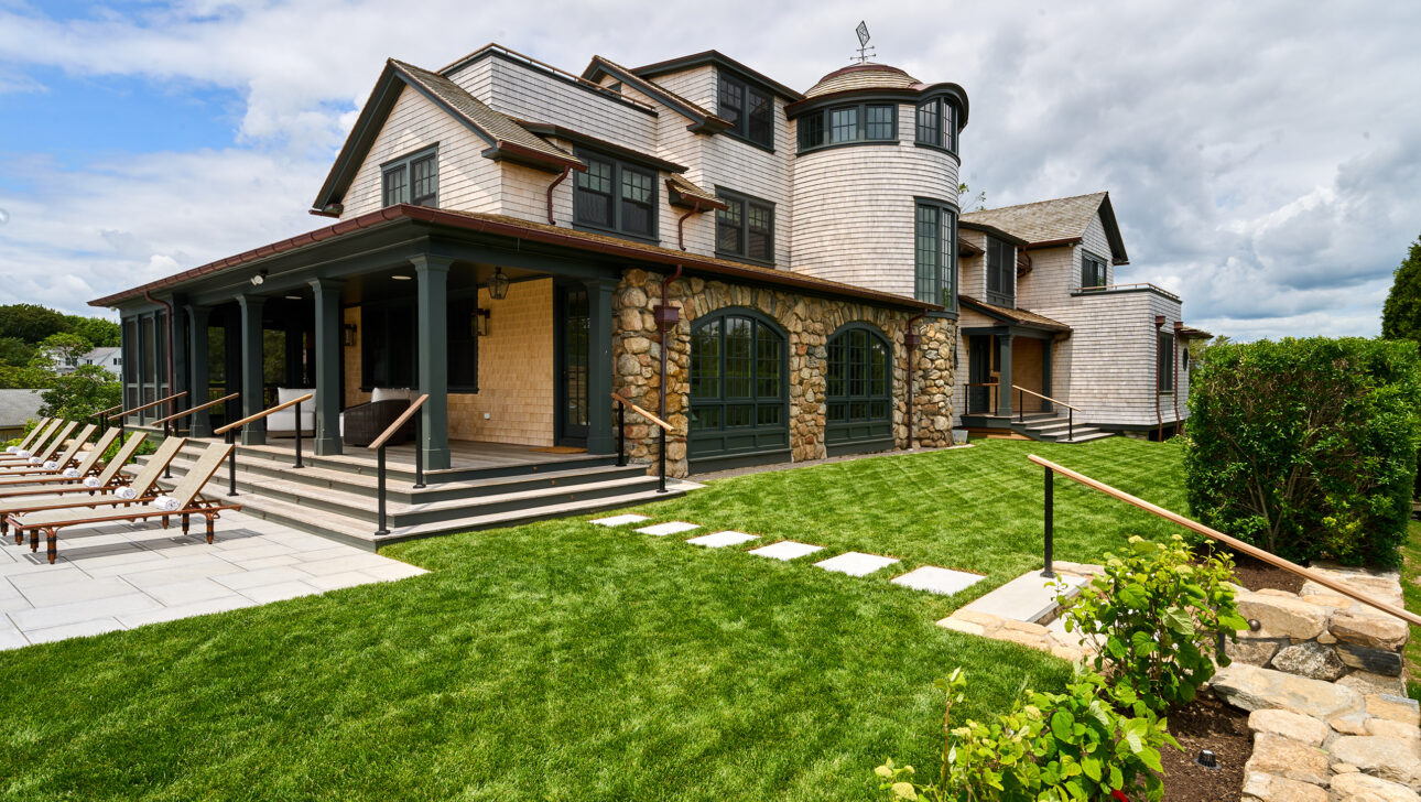 A large stone and wood house with a grassy lawn, patio with lounge chairs, and cloudy sky in the background.