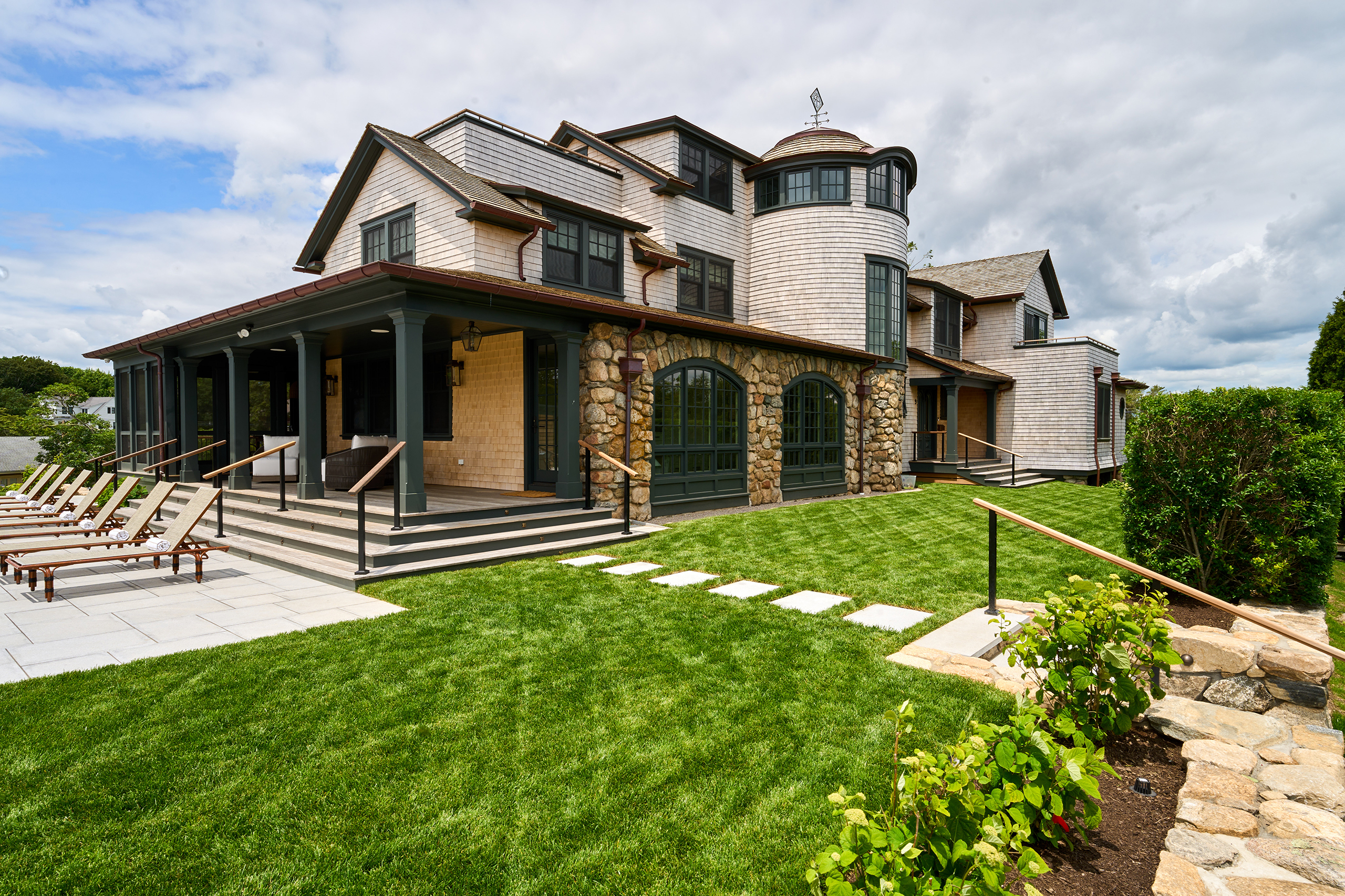 A large stone and wood house with a grassy lawn, patio with lounge chairs, and cloudy sky in the background.