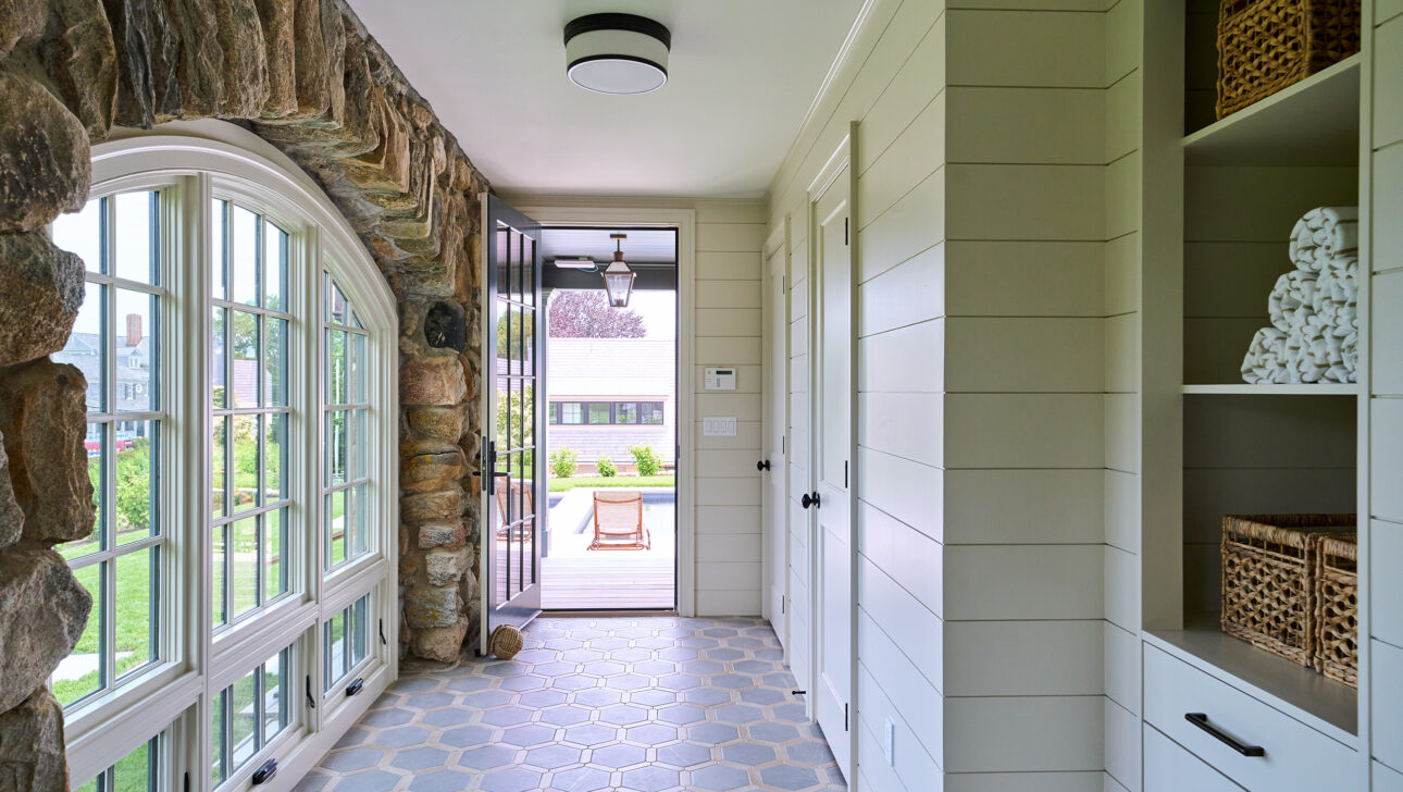 A hallway with large stone arches and hexagonal floor tiles. The space leads to an open door, showing a view of a patio and lawn outside. Shelving on the right holds towels.