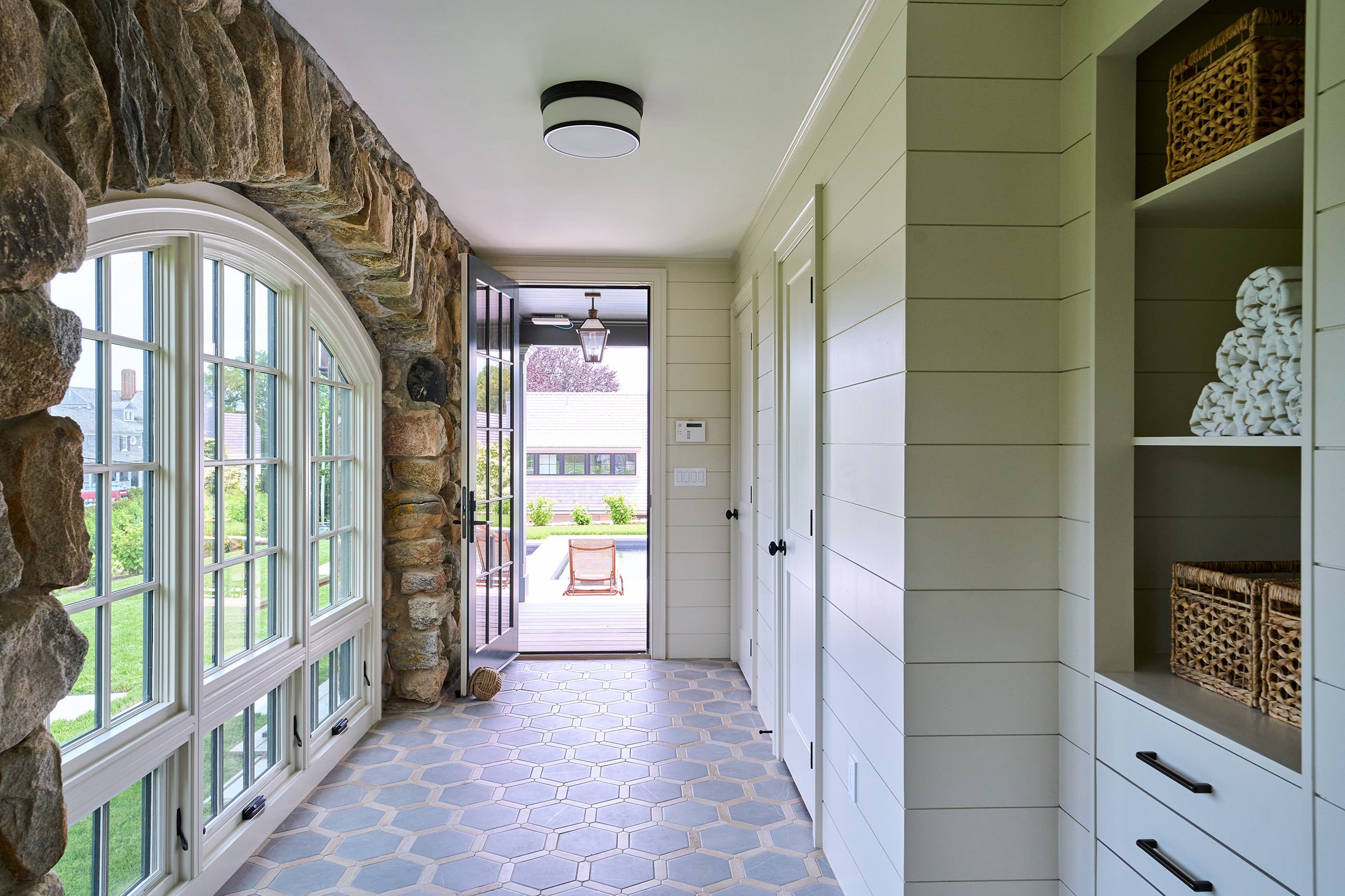 A hallway with large stone arches and hexagonal floor tiles. The space leads to an open door, showing a view of a patio and lawn outside. Shelving on the right holds towels.