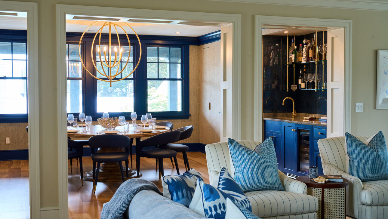 A modern open-plan living space with a dining room featuring a round table and a chandelier. Blue cabinetry is visible in the bar area. The seating area has striped chairs and patterned pillows.