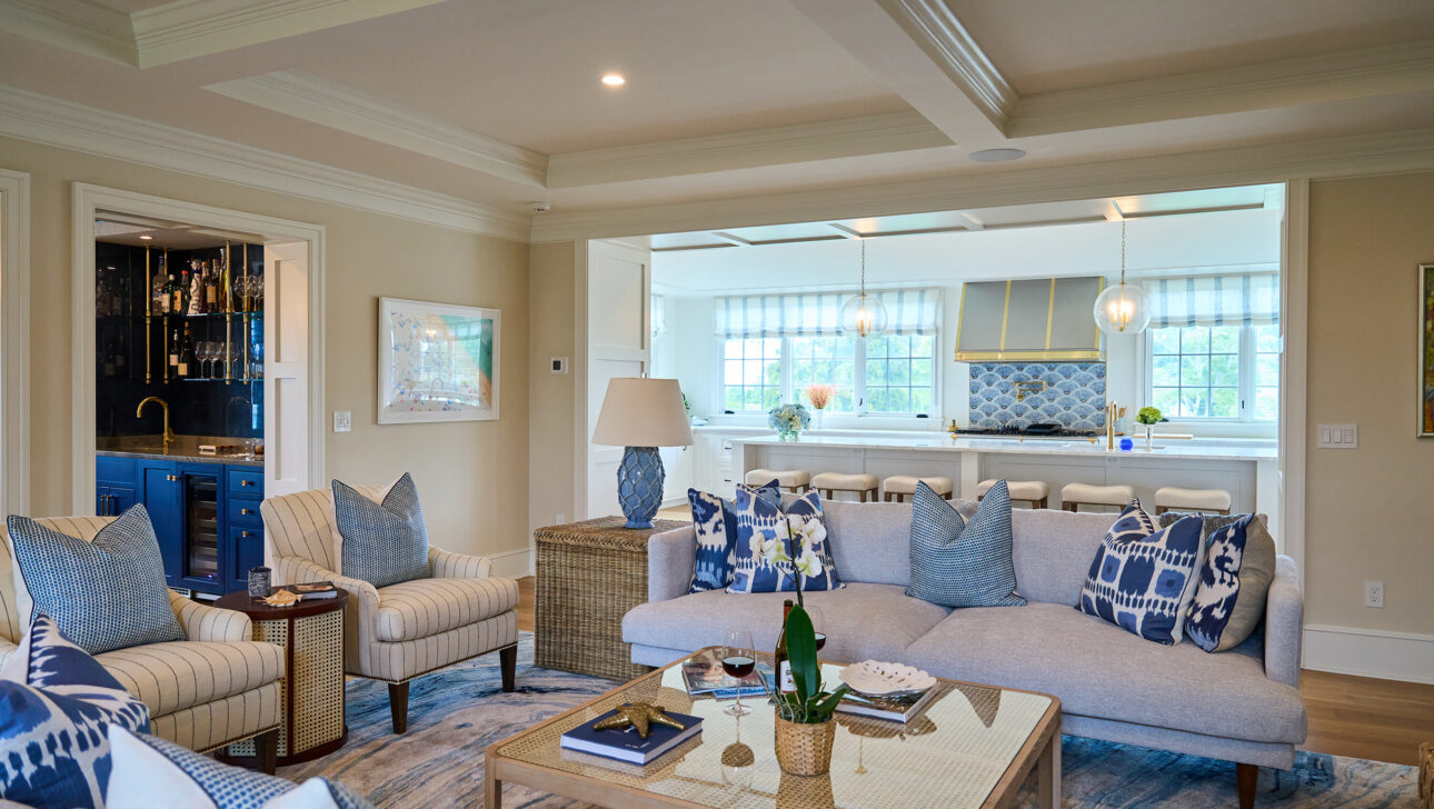 Modern living room with beige sofas, blue and white pillows, and a wicker coffee table. The space connects to a bright kitchen with blue accents and large windows.