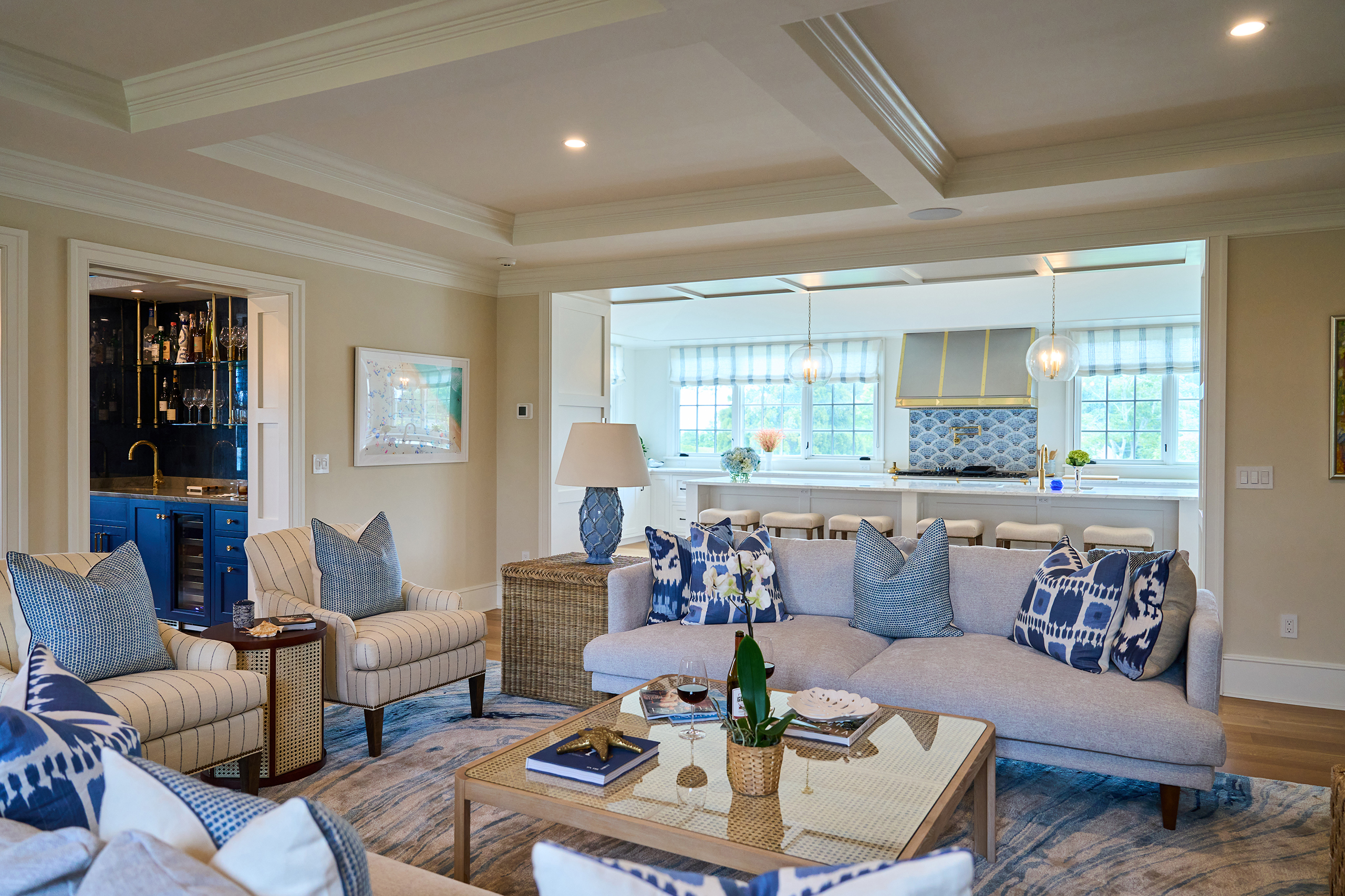 Modern living room with beige sofas, blue and white pillows, and a wicker coffee table. The space connects to a bright kitchen with blue accents and large windows.