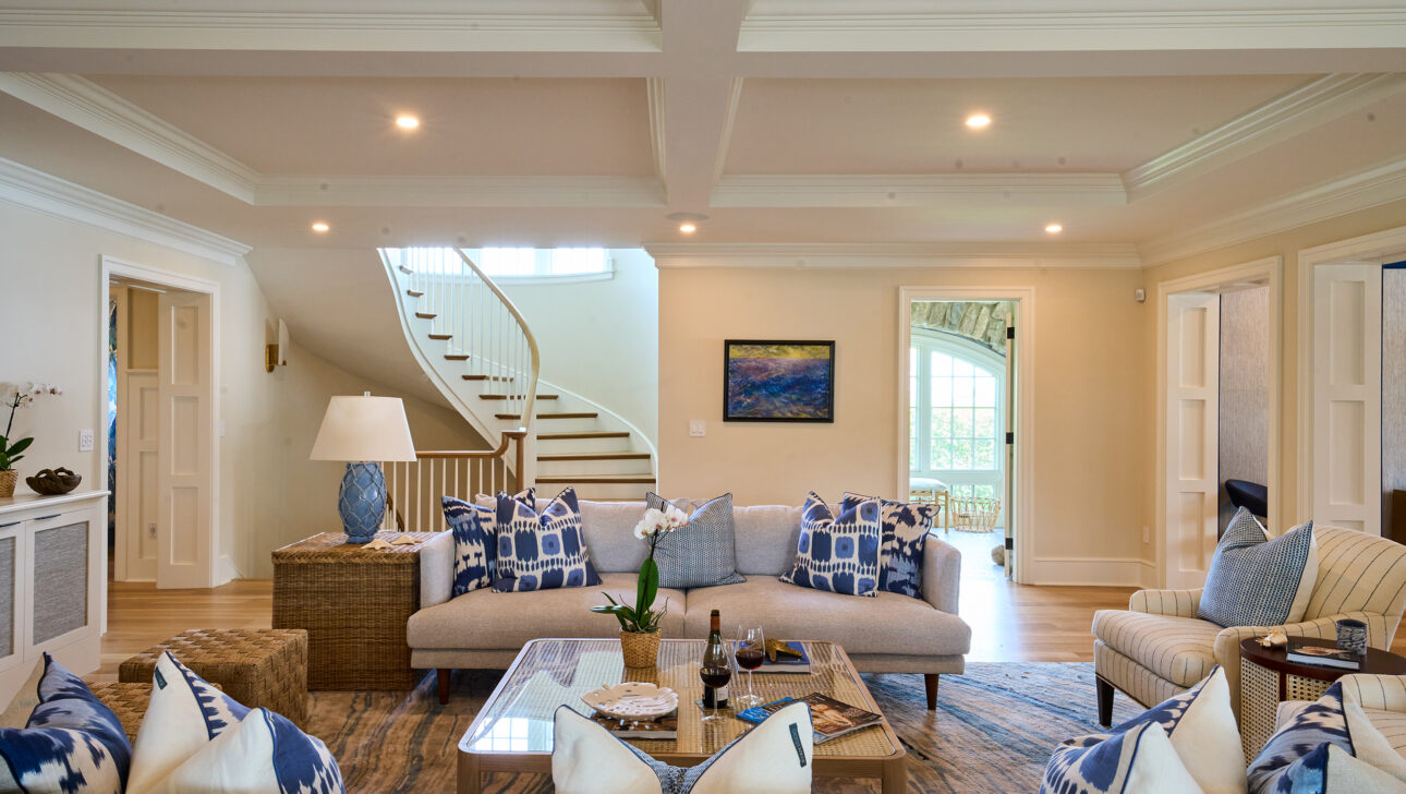 A spacious living room with two sofas, patterned cushions, a coffee table, and a staircase in the background. Neutral tones and soft lighting emphasize a relaxed, elegant atmosphere.