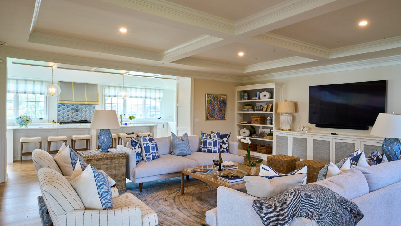Spacious living room with beige sofas, blue pillows, a large TV, and an open view into a bright kitchen with bar stools.