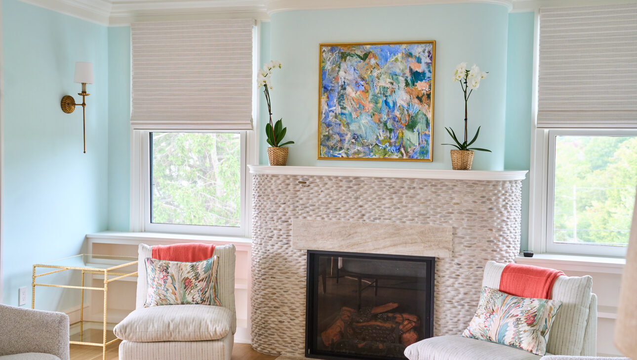 A living room with a light blue wall, a textured fireplace, and a colorful painting. Two chairs with floral pillows flank the fireplace, and windows on either side have shades.