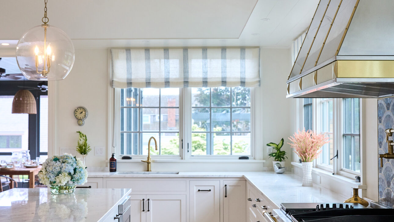 A modern kitchen with white cabinets, marble countertops, and a large window. Brass fixtures, a pendant light, and a range hood accent the space. A vase with flowers decorates the counter.
