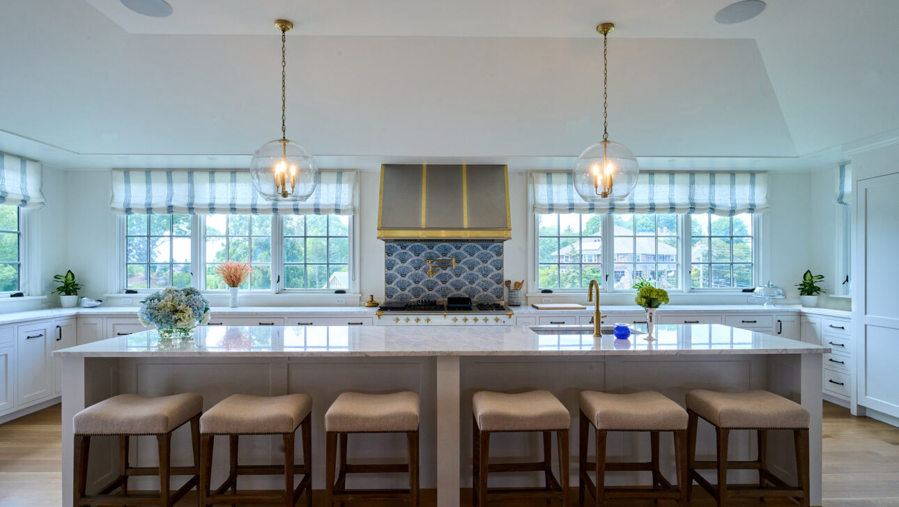 Spacious kitchen with a large island featuring six stools. Two pendant lights hang above, and striped curtains adorn the windows. White cabinets and a blue accented range hood complete the scene.