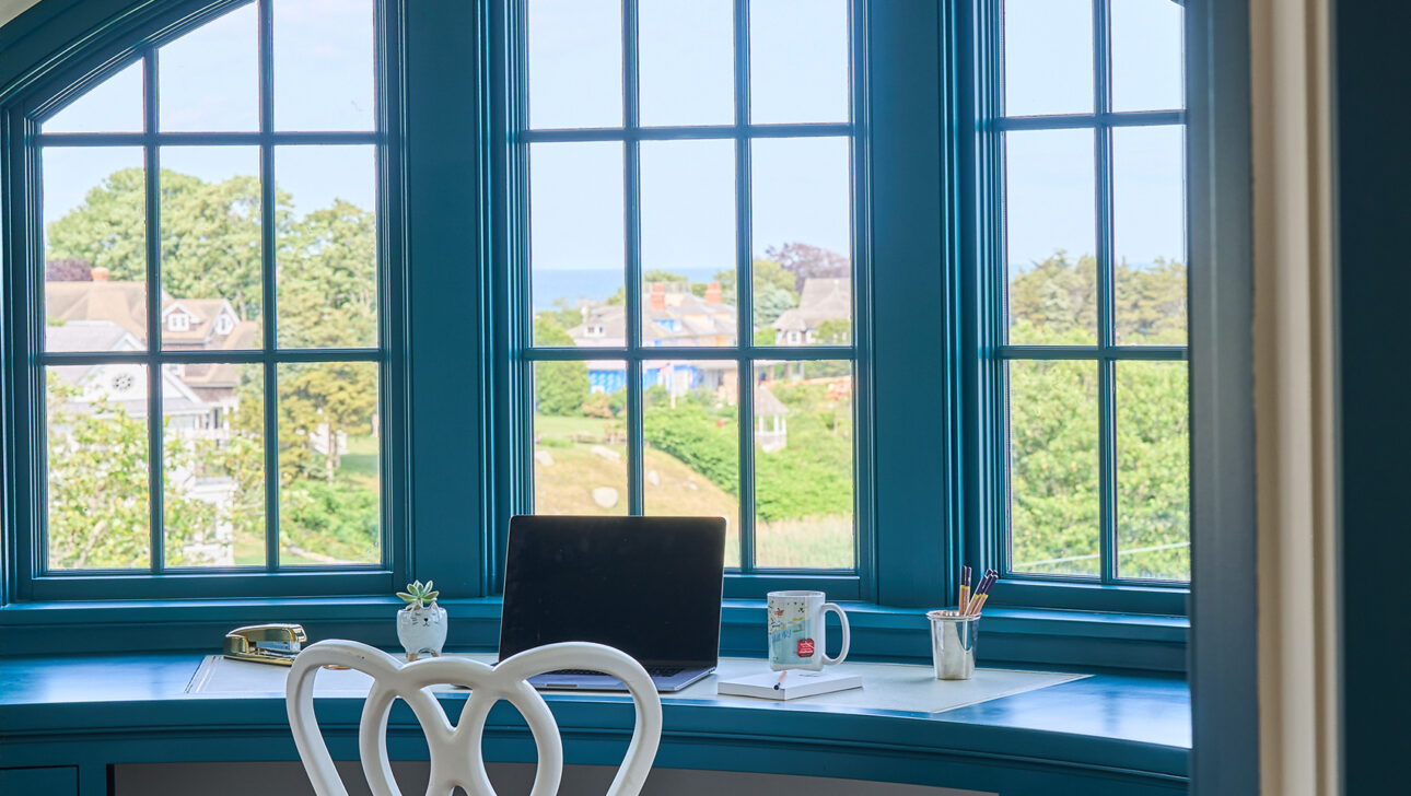 A home office with a blue curved desk by a large arched window, a white chair, a laptop, a cup, and stationery. The window offers a view of trees and buildings in the distance.