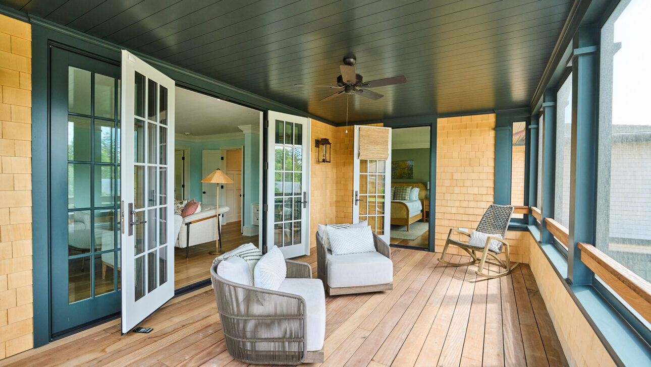 A sunlit enclosed porch with wooden flooring, two cushioned chairs, a rocking chair, and open French doors leading to an interior room. A ceiling fan is mounted on the dark ceiling.