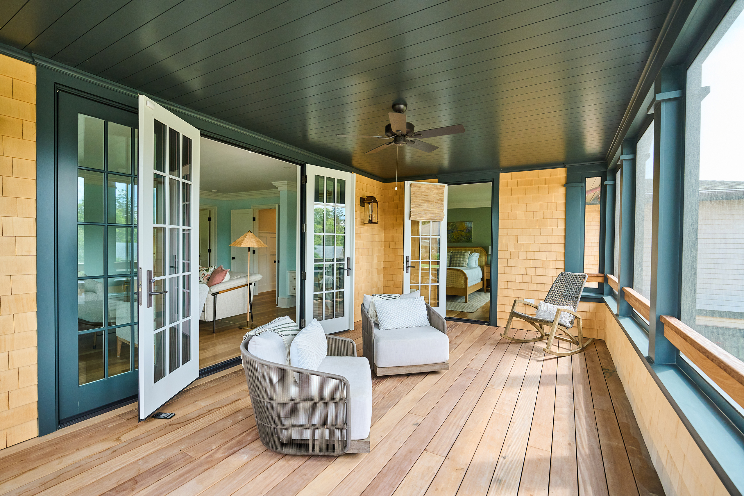 A sunlit enclosed porch with wooden flooring, two cushioned chairs, a rocking chair, and open French doors leading to an interior room. A ceiling fan is mounted on the dark ceiling.