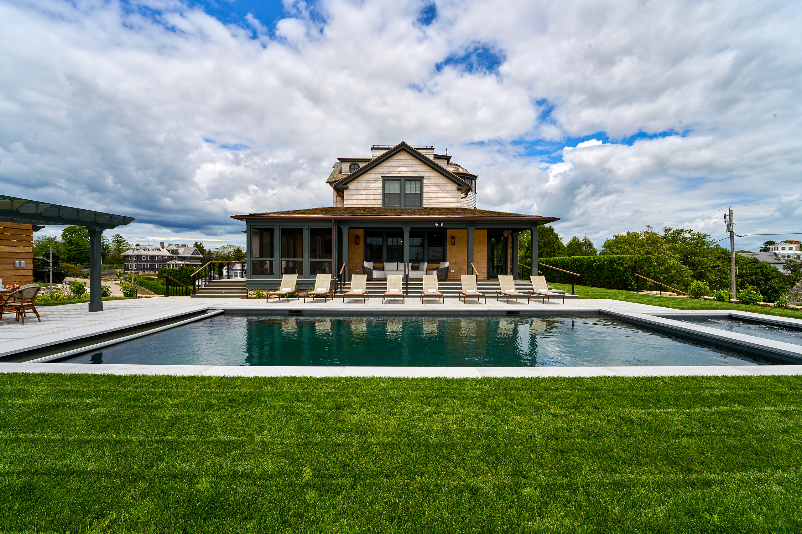 A large house with a wooden exterior is flanked by an outdoor swimming pool and several lounge chairs, set against a cloudy sky backdrop.