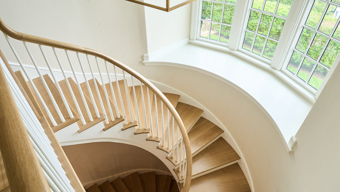 A spiral wooden staircase with white railings curves downwards. A large window with multiple panes is on the right, and a geometric chandelier hangs above.