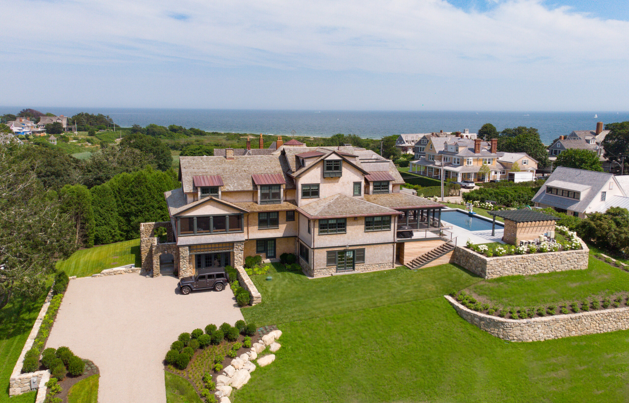 Aerial view of a large seaside house with a pool, a driveway with a parked car, and landscaped lawns, overlooking the ocean.