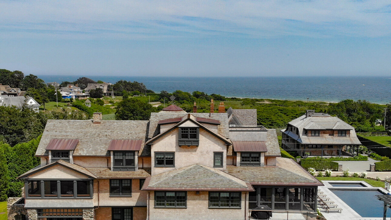 A large coastal house with multiple levels, a wraparound porch, and a black car parked outside, situated near the ocean with a swimming pool in the backyard.