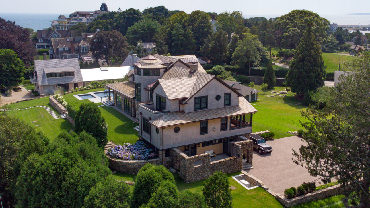 Aerial view of a large house with a stone facade, surrounded by lush greenery and a garden, with additional buildings and a pool visible in the background.