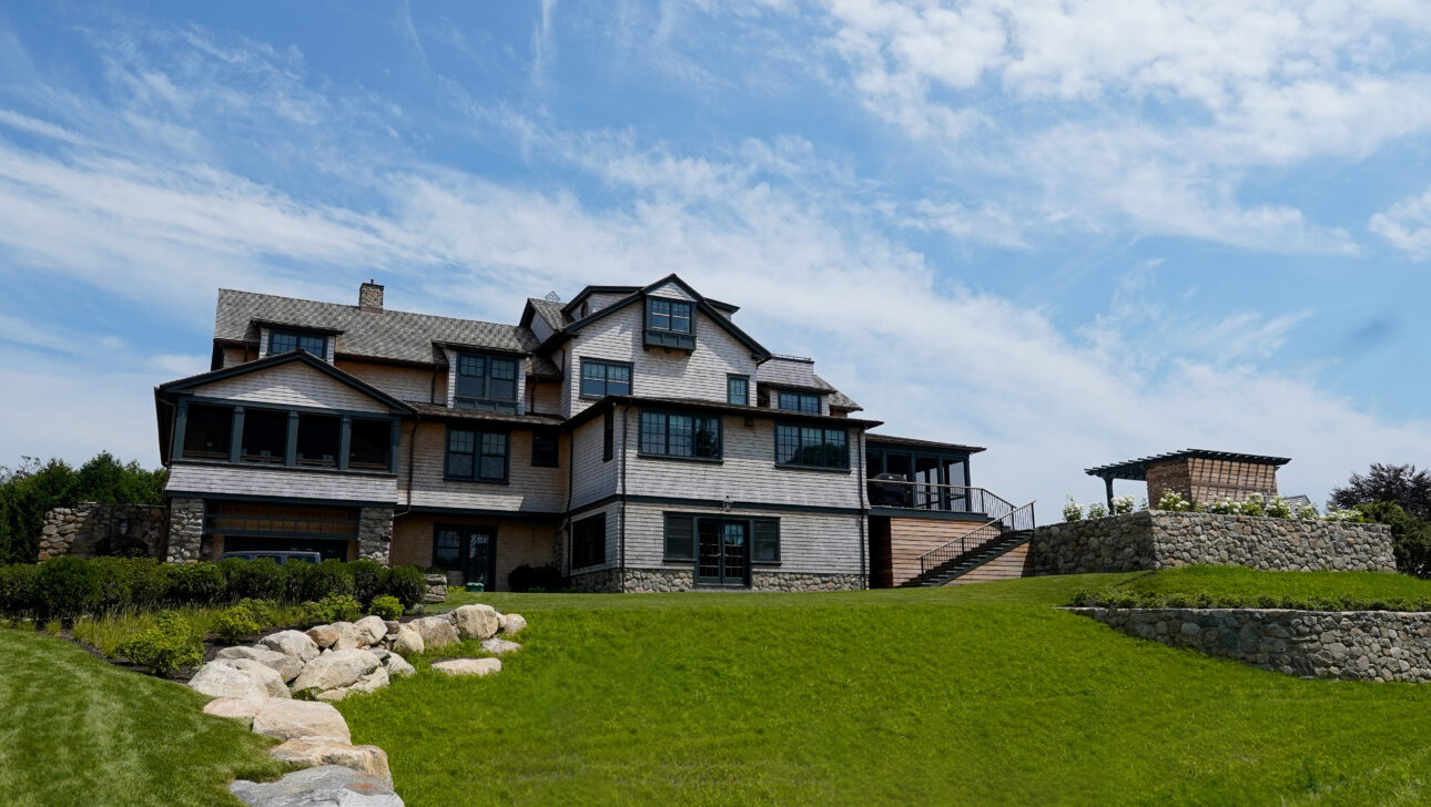 Large multi-story house with wooden siding and stone accents, situated on a grassy hill under a cloudy sky.