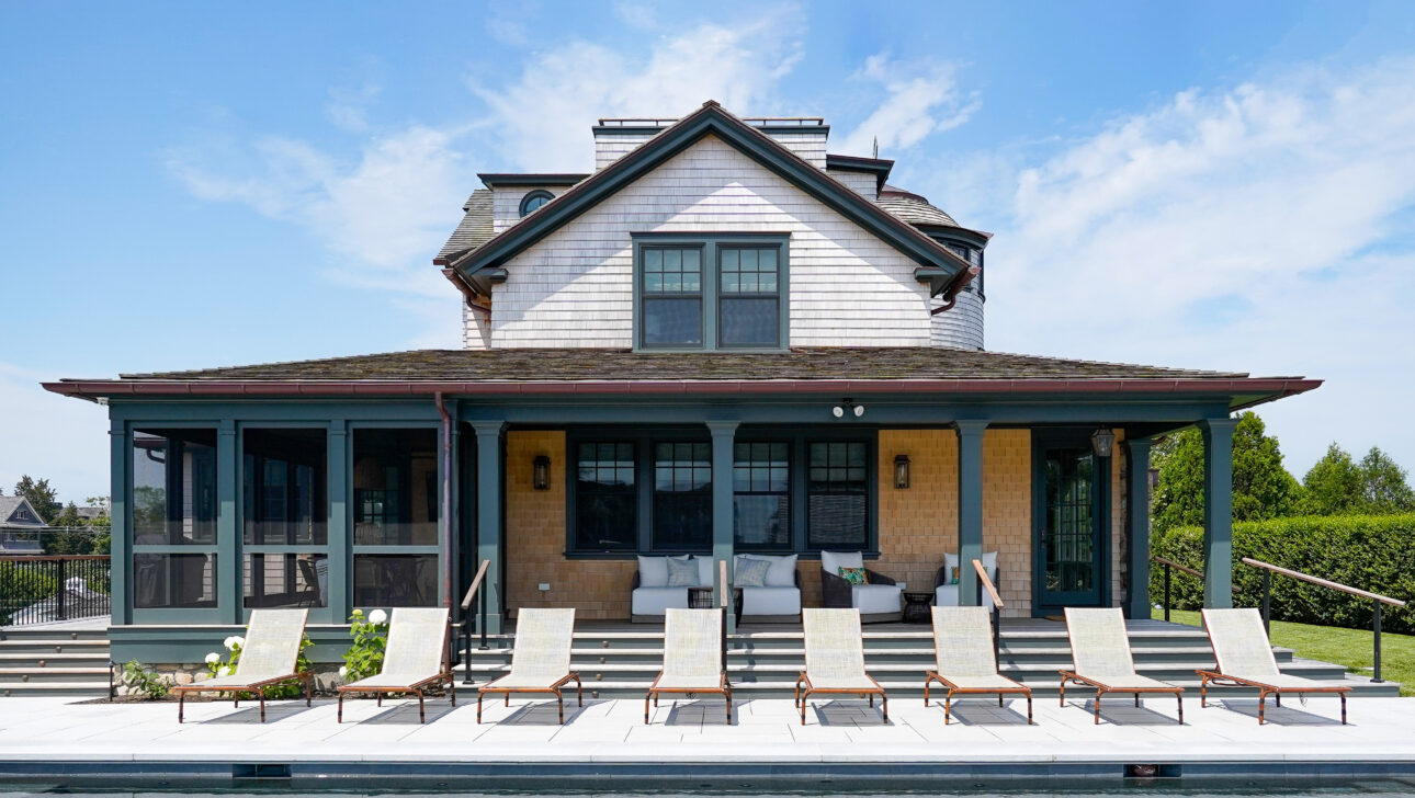 A large house with a gabled roof and wide veranda overlooks a rectangular swimming pool, with several lounge chairs aligned on the poolside patio.