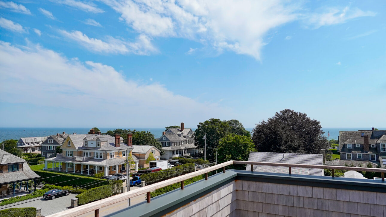 Rooftop view of a coastal neighborhood with houses, trees, and the ocean under a partly cloudy sky.