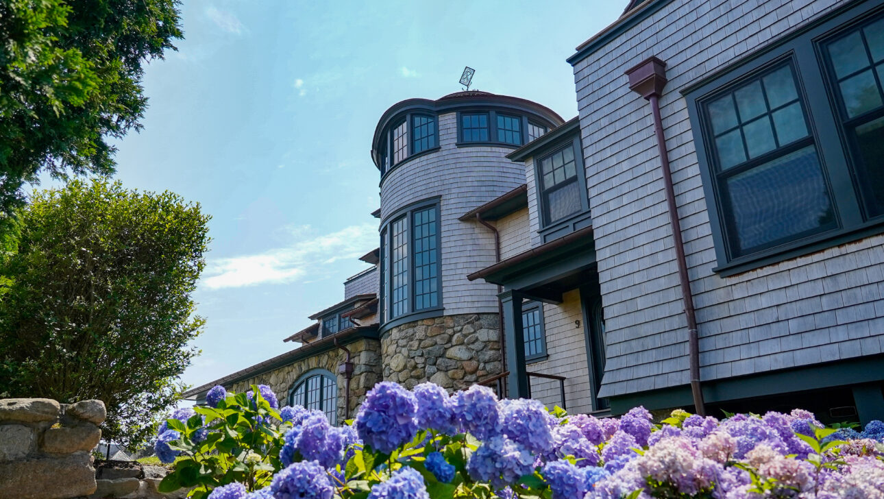 Large stone and shingle house with a round tower, surrounded by blooming purple and blue hydrangeas under a clear blue sky.