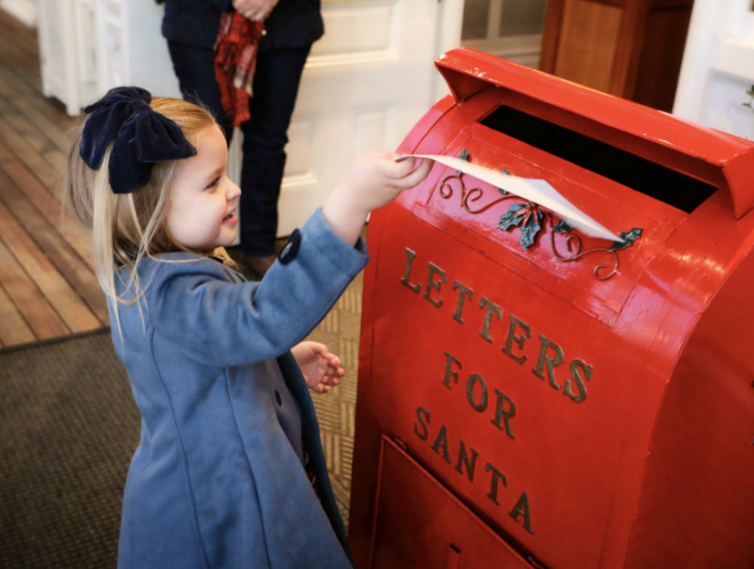 Young girl in a blue coat places a letter in a red mailbox labeled "Letters for Santa.