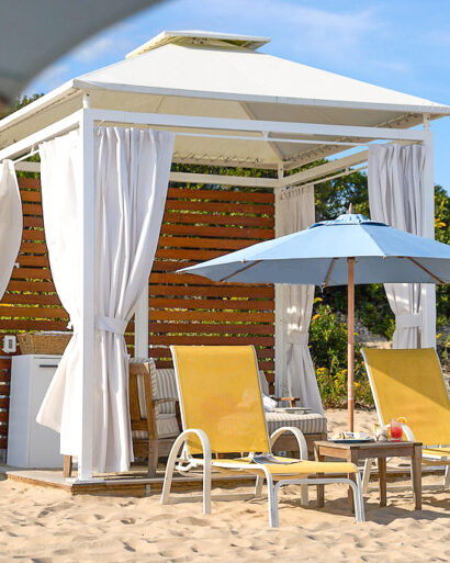 A beach cabana with white curtains, two yellow lounge chairs, and a blue umbrella on sandy ground near greenery.