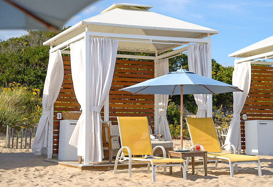 A beach cabana with white curtains, two yellow lounge chairs, and a blue umbrella on sandy ground near greenery.