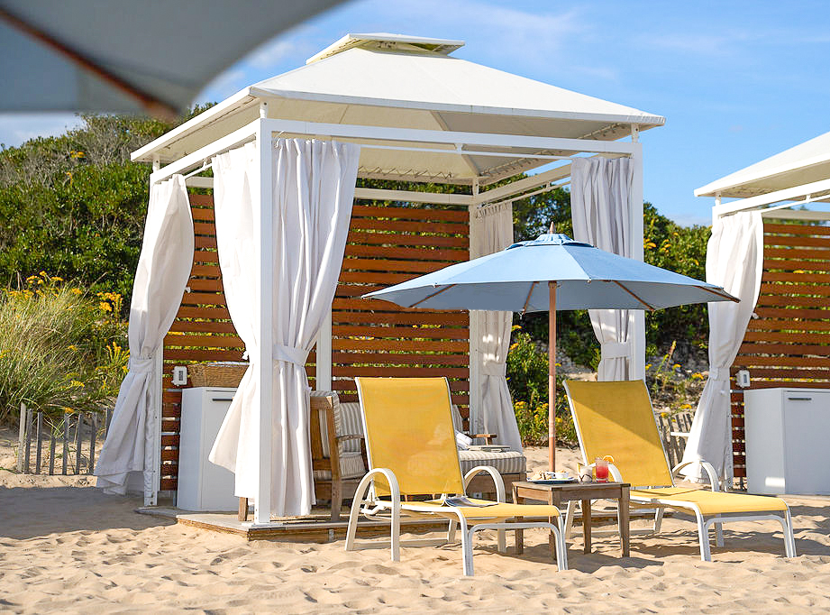 A beach cabana with white curtains, two yellow lounge chairs, and a blue umbrella on sandy ground near greenery.