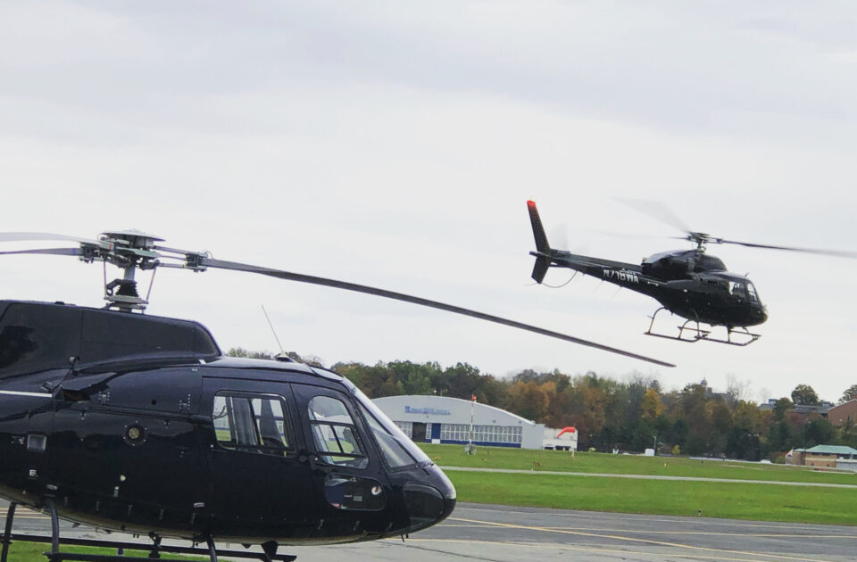 Two black helicopters at a small airfield: one is on the ground, and the other is flying. Overcast sky and grassy area in the background.